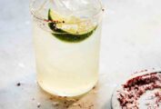 A highball filled with a long margarita, ice, a lime wedge, and a dish of chili salt for rimming on the side