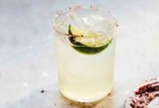 A highball filled with a long margarita, ice, a lime wedge, and a dish of chili salt for rimming on the side