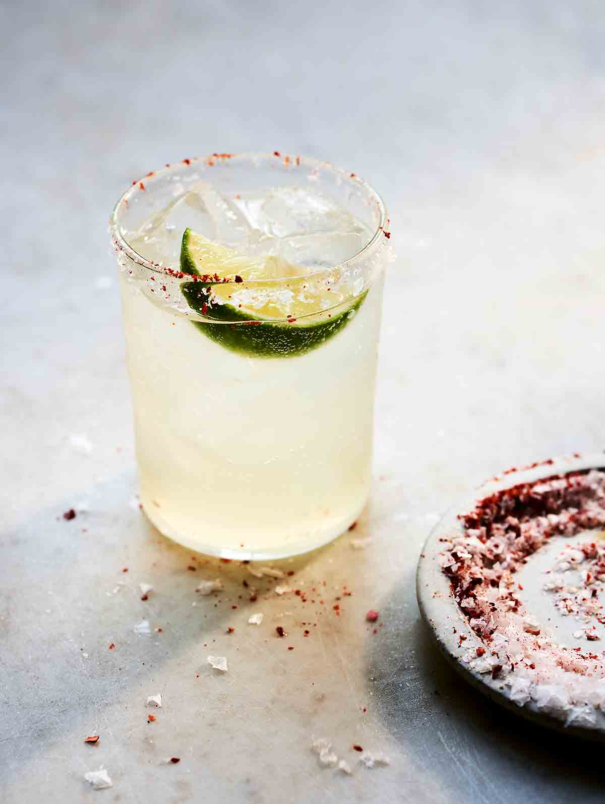 A highball filled with a long margarita, ice, a lime wedge, and a dish of chili salt for rimming on the side