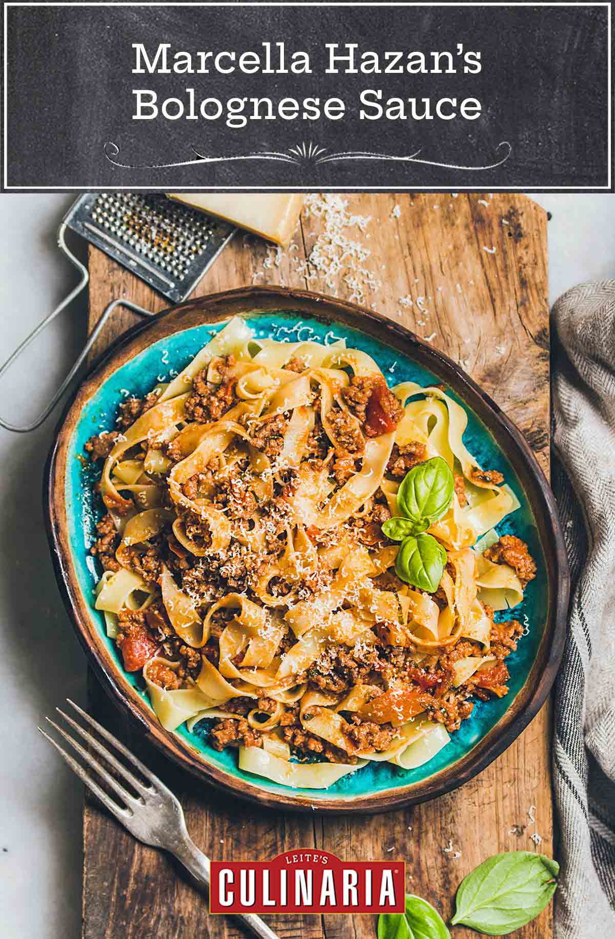 A blue bowl filled with pappardelle noodles and Marcella Hazan's bolognese sauce on a wooden board with a block of Parmesan and a grater beside the bowl.
