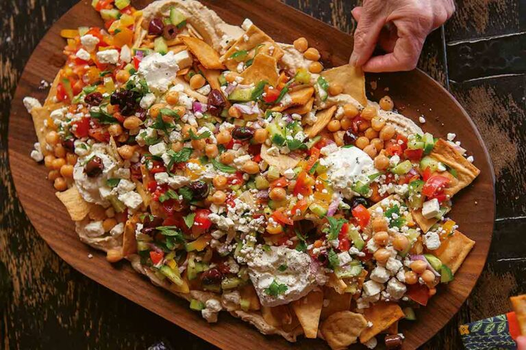 A platter of Mediterranean nachos topped with yogurt, hummus, cucumbers, tomatoes, olives, feta, and chickpeas on a wooden platter