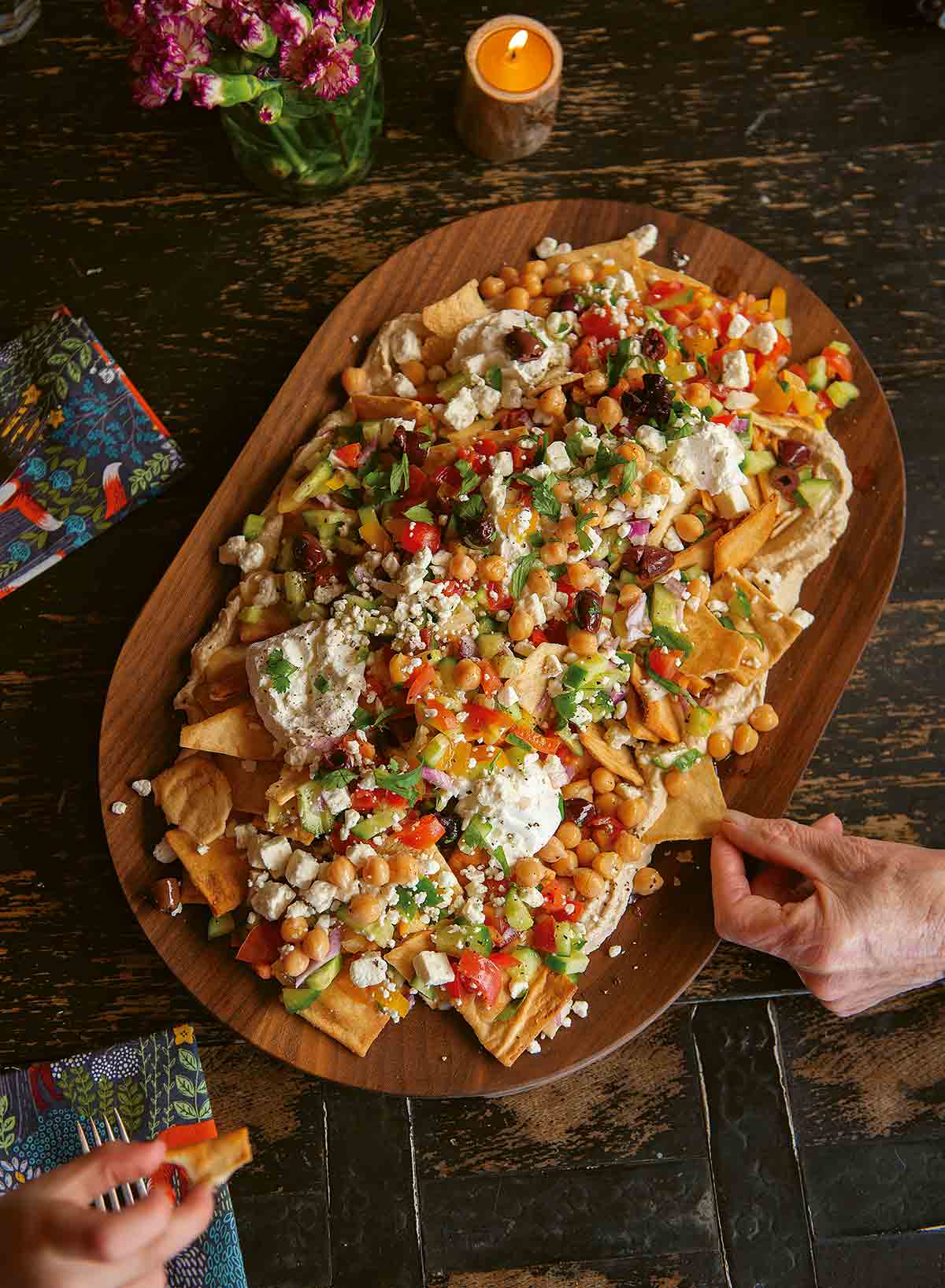 A platter of Mediterranean nachos topped with yogurt, hummus, cucumbers, tomatoes, olives, feta, and chickpeas on a wooden platter