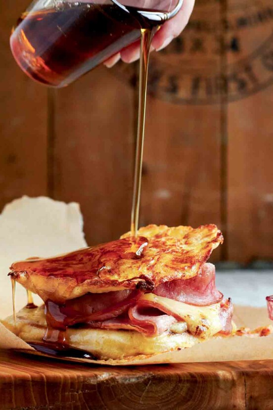 Maple syrup being poured over a monte cristo sandwich that is on a piece of parchment paper.