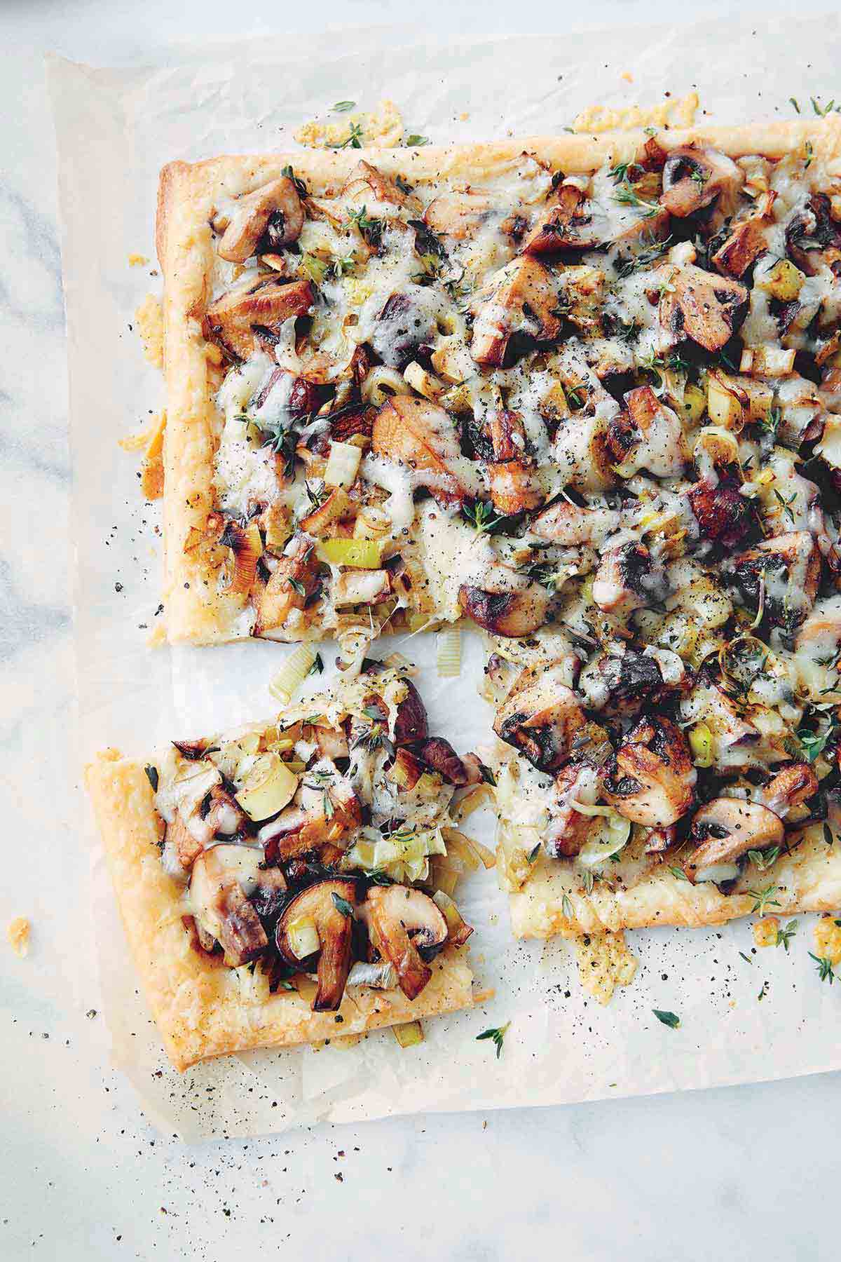 A sheet of parchment paper with a rectangular mushroom, leek, and Gruyere tart with a square piece cut from the corner, and garnished with thyme.
