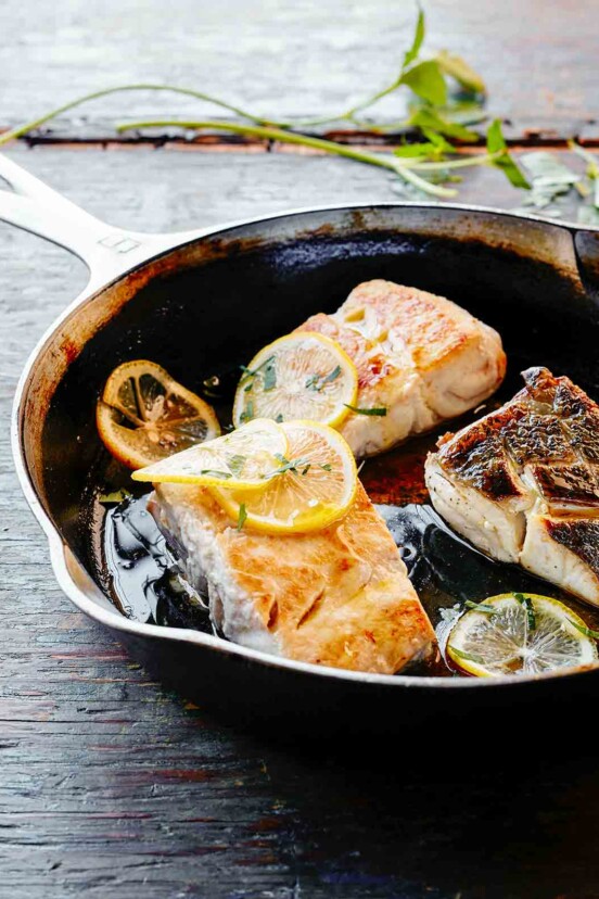 A skillet with three pieces of pan fried fish and lemon slices