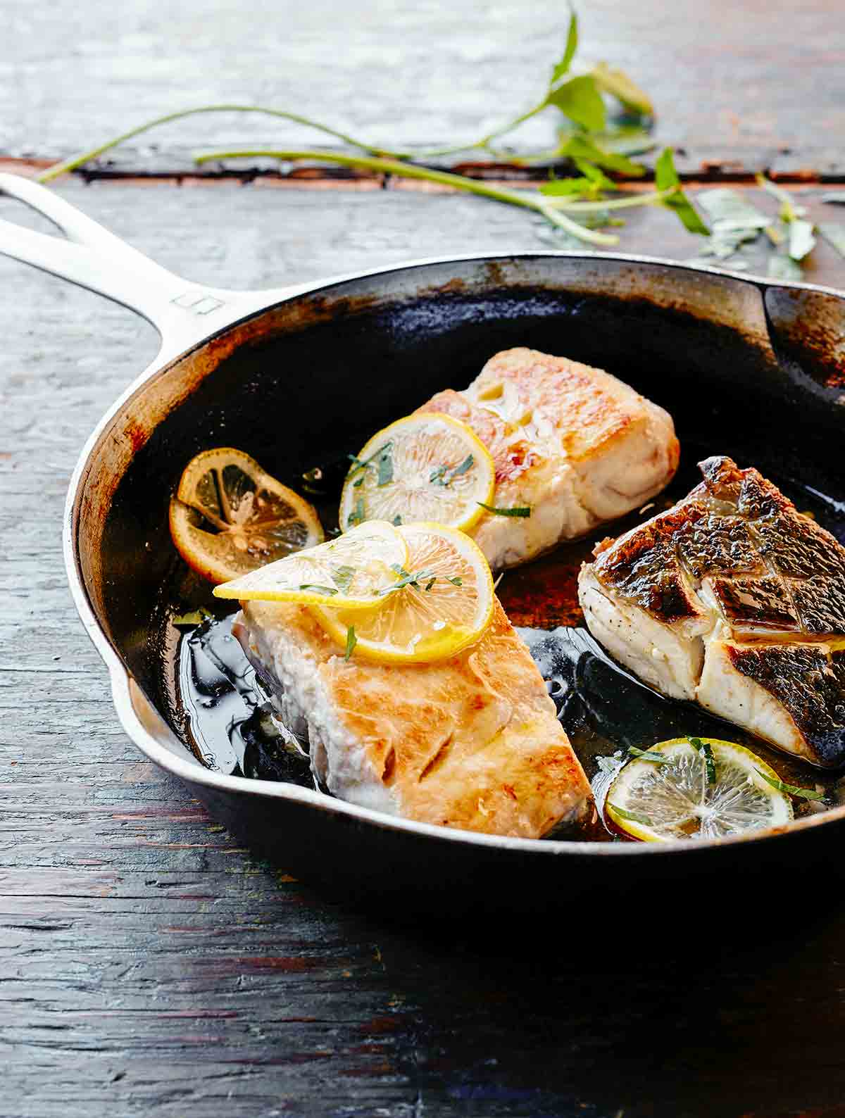 A skillet with three pieces of pan fried fish and lemon slices