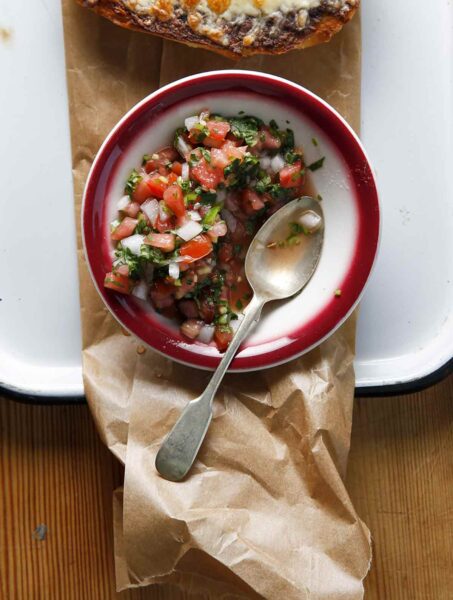A bowl of pico de gallo with a spoon resting inside set on a piece of parchment in a rimmed baking tray.
