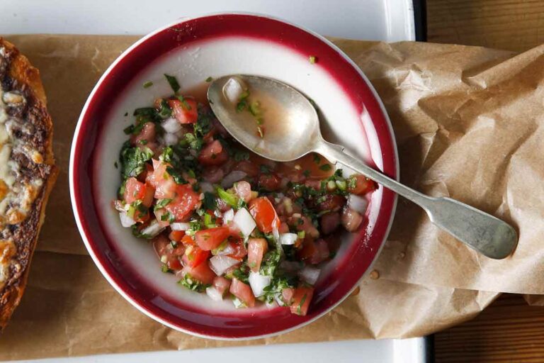 A bowl of pico de gallo with a spoon resting inside set on a piece of parchment in a rimmed baking tray.