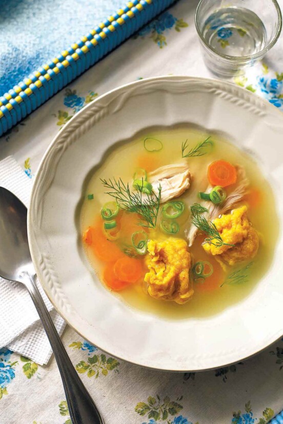 A bowl of Ukrainian chicken dumpling soup with shredded chicken, dill, scallions, and carrots and a spoon on the side