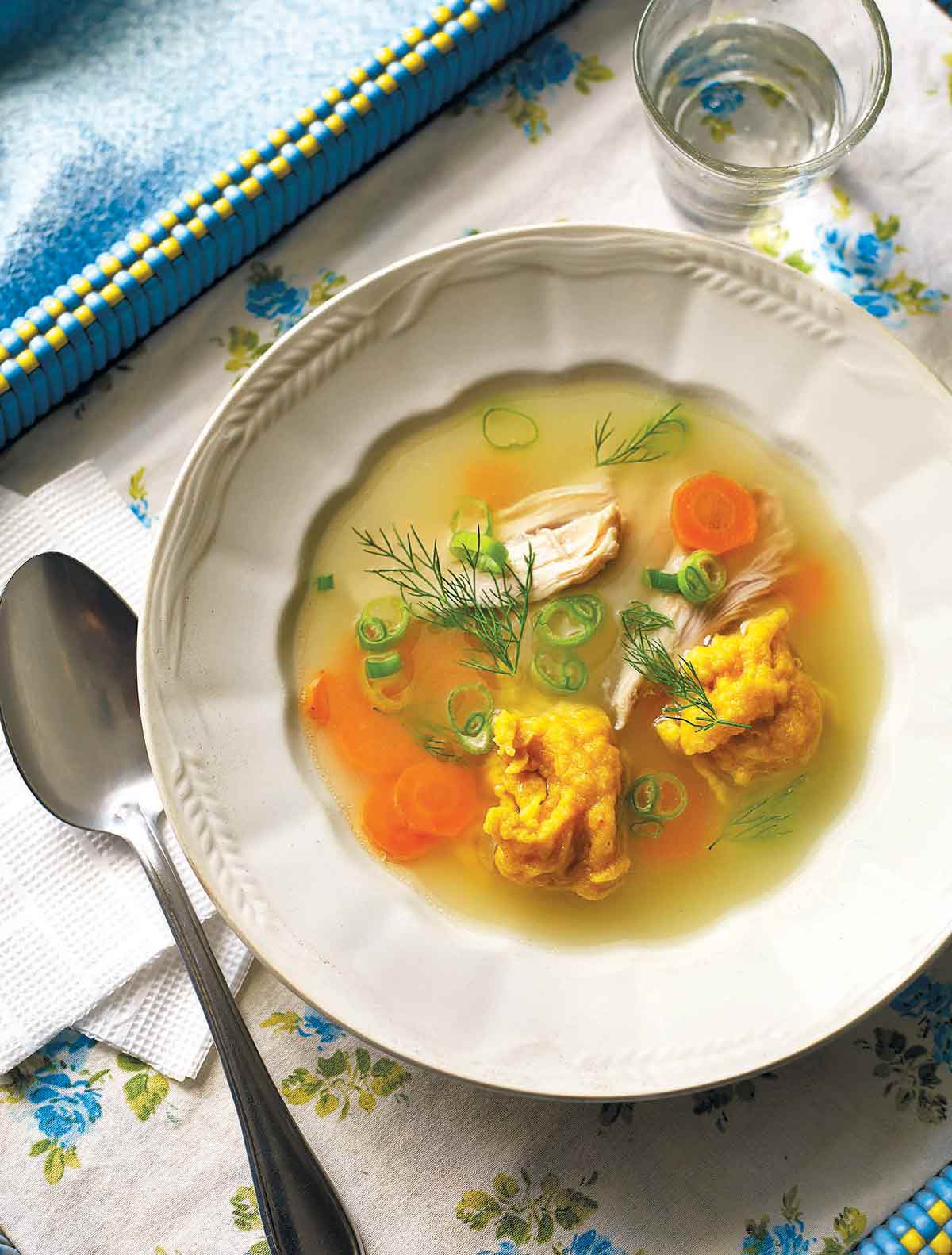 A bowl of Ukrainian chicken dumpling soup with shredded chicken, dill, scallions, and carrots and a spoon on the side