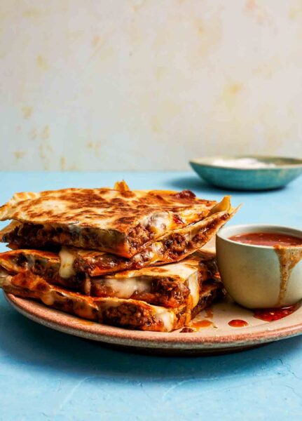 Four pieces of cheesy beef and caramelized onion quesadillas stacked on a plate with a bowl of dipping sauce on the side.