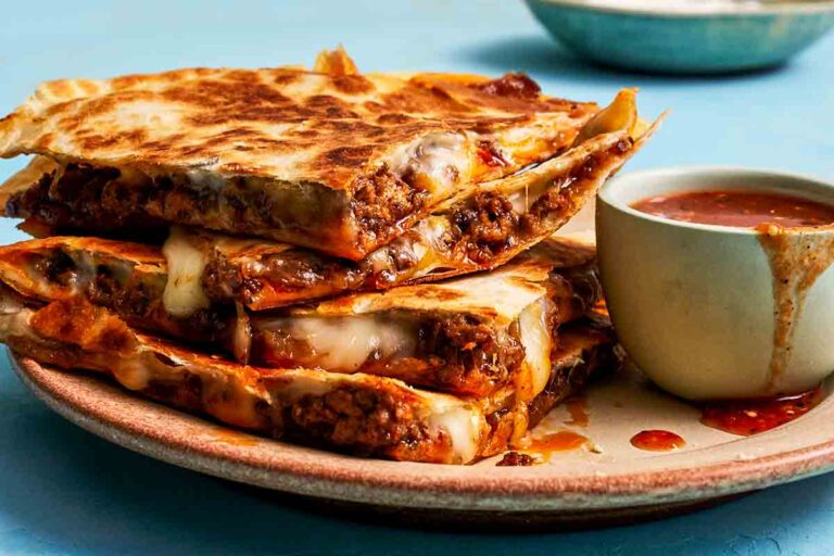Four pieces of cheesy beef and caramelized onion quesadillas stacked on a plate with a bowl of dipping sauce on the side.