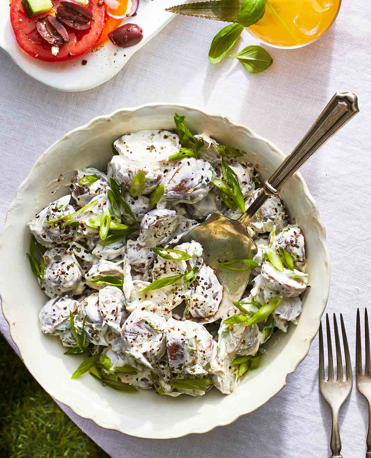 A white porcelain bowl filled with creamy red potato salad, topped with sliced scallions and a serving spoon resting inside