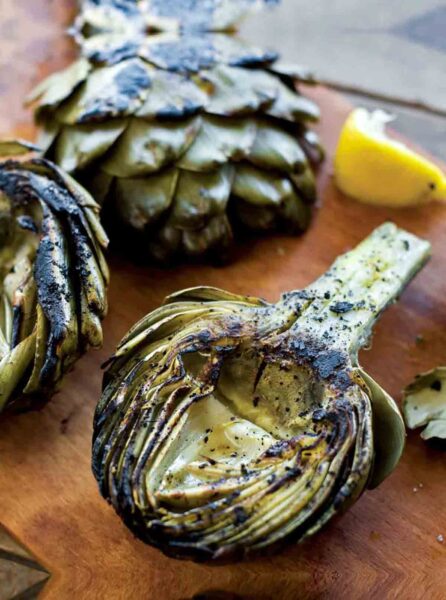 Three grilled artichoke halves, some lemon wedges, and dip on a wooden peel