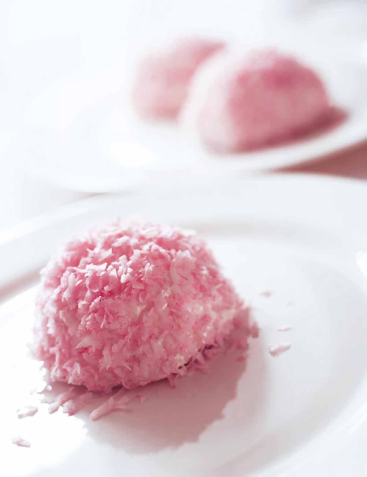 A pink homemade sno ball--coconut-covered dome--on a white plate with two more sno balls in the back.
