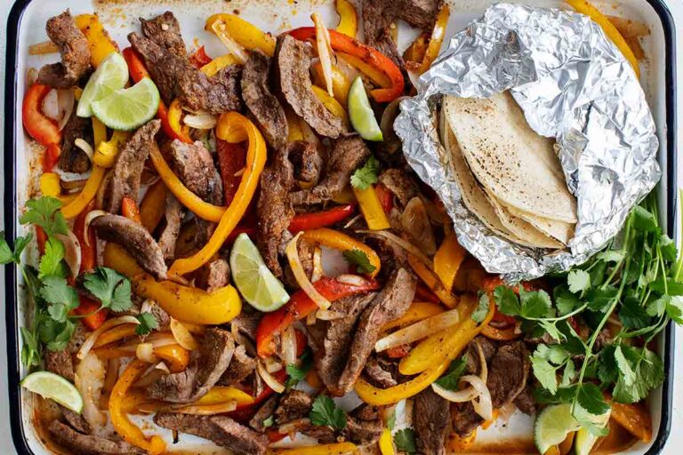 A metal tray filled with the fillings for sheet pan steak fajitas -- sliced steak, peppers, lime wedges, tortillas, and cilantro