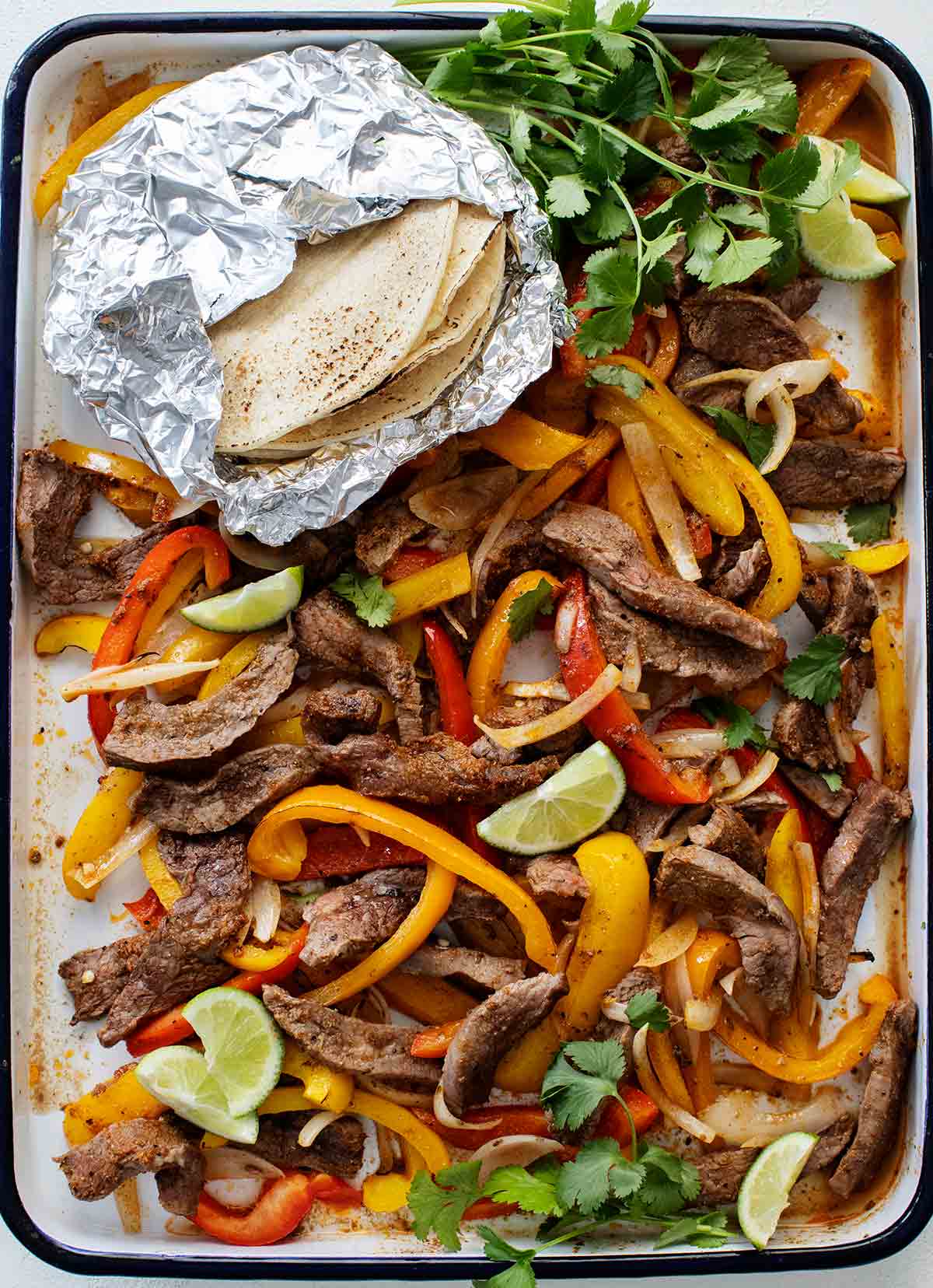 A metal tray filled with the fillings for sheet pan steak fajitas -- sliced steak, peppers, lime wedges, tortillas, and cilantro