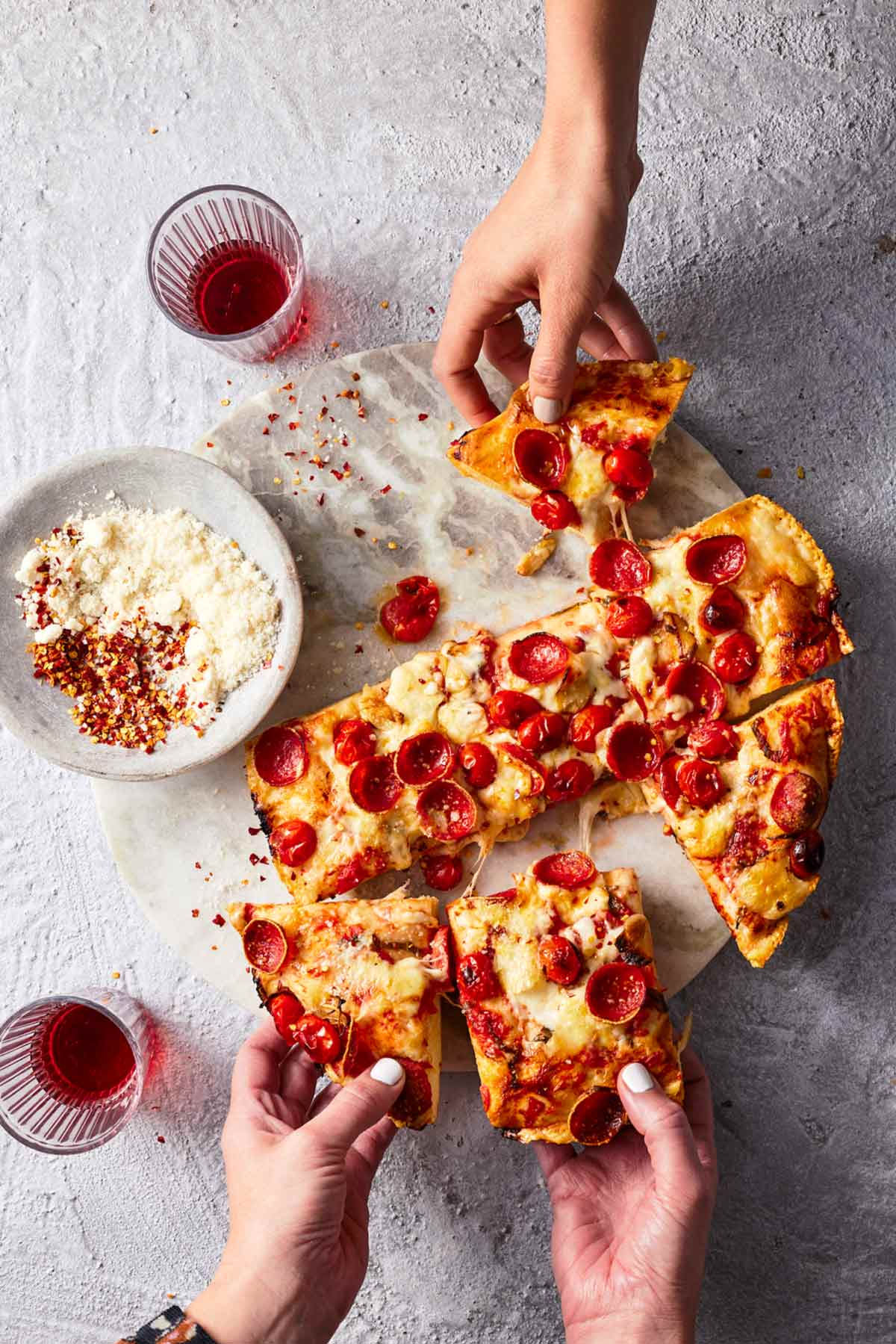 Händer som tar tag i bitar av stekpanna pepperoni pizza med två glas på sidan och en skål med parmesan och pepparflingor