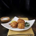 Three Spanish cod fritters on a square white plate with parsley on a yellow napkin and black tray
