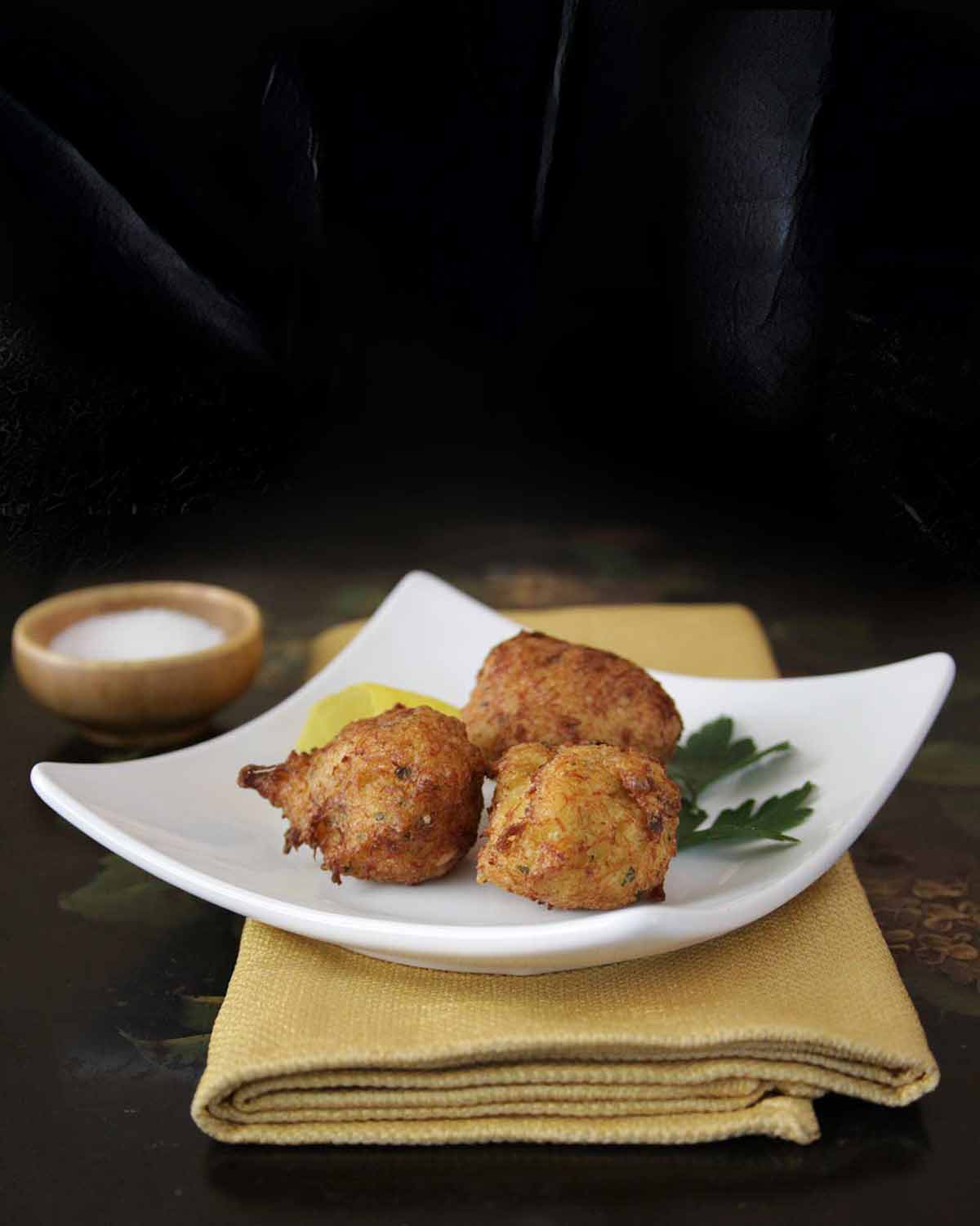 Three Spanish cod fritters, aka buñuelos de bacalao, on a square white plate with parsley on a yellow napkin and black tray