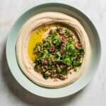 A pale blue bowl filled with white bean hummus, topped with parsley, olives, seeds, and oil
