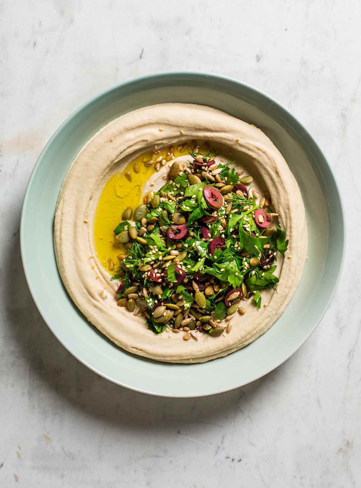 A pale blue bowl filled with white bean hummus, topped with parsley, olives, seeds, and oil