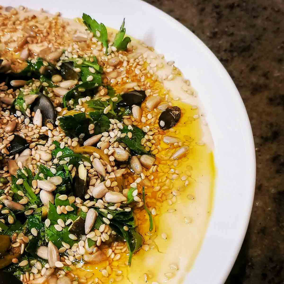 A white bowl of white bean hummus topped with seeds, parsley, olives, and oil