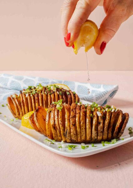 Two air fryer hasselback potatoes on a white plate with a person squeezing lemon over the top.