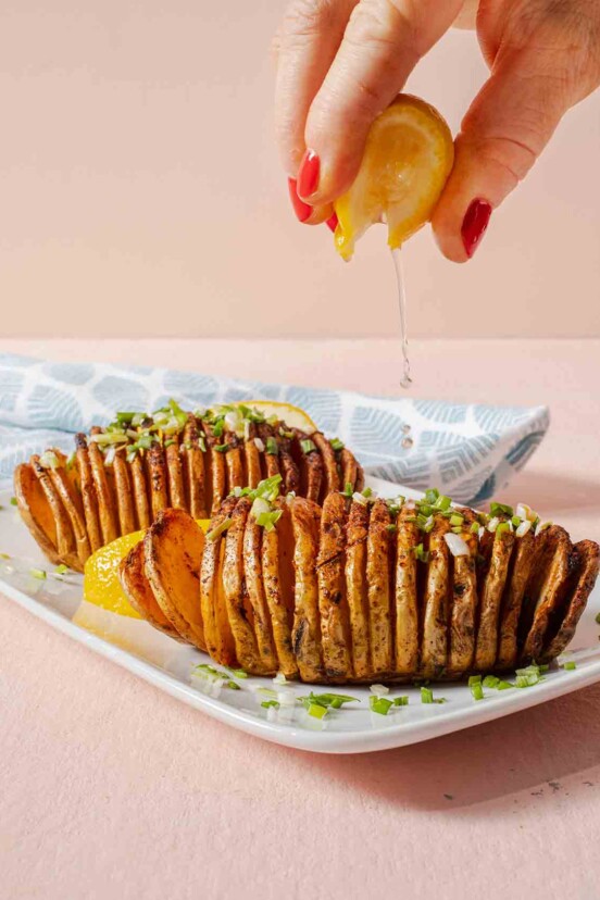 Two air fryer hasselback potatoes on a white plate with a person squeezing lemon over the top.