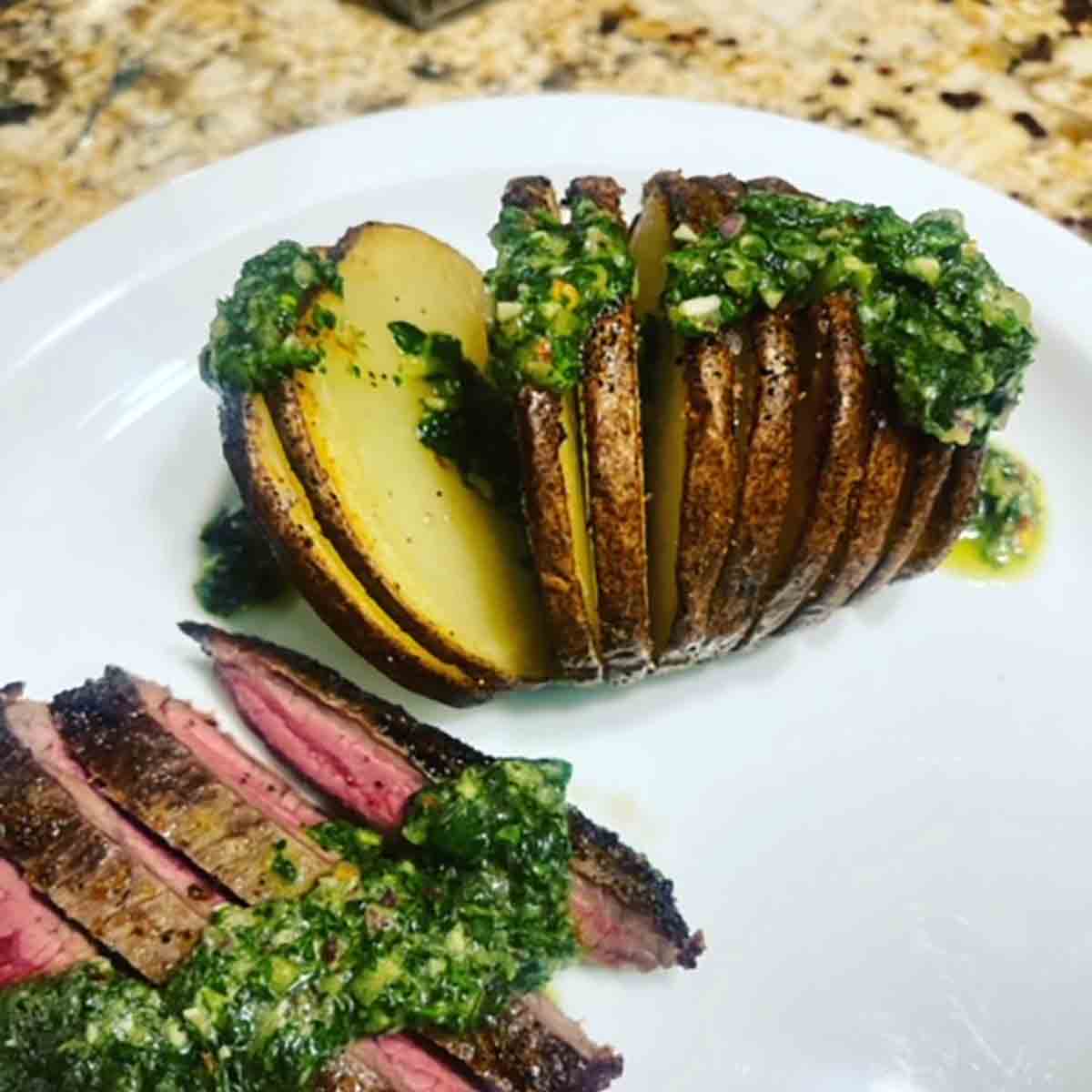 An air fryer hasselback potato and a sliced steak with herb sauce on top on a white plate.