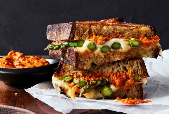 Two halves of a toasted asparagus and cheese sandwich stacked on top of each other on a sheet of parchment with a bowl of Romesco sauce on the side.