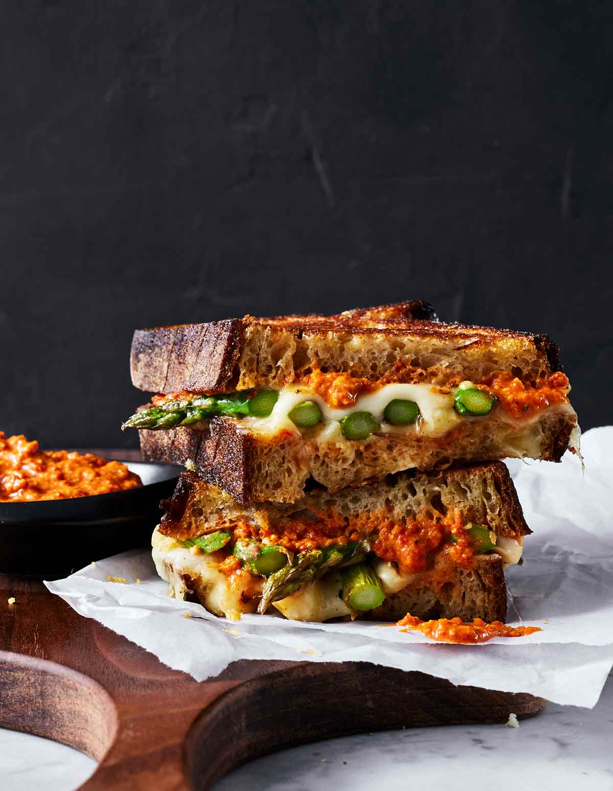 Two halves of a toasted asparagus and cheese sandwich stacked on top of each other on a sheet of parchment with a bowl of Romesco sauce on the side.