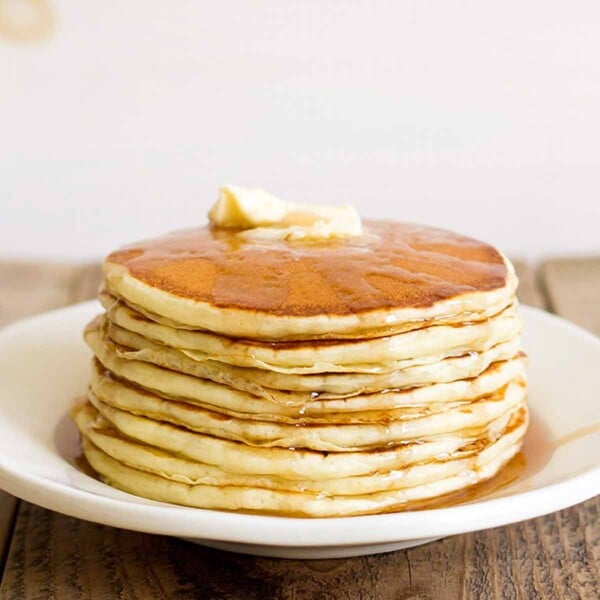 A stack of fluffy buttermilk pancakes on a white plate with syrup and butter on top.