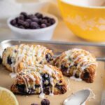 Three glazed blueberry lemon scones on a parchment-lined baking sheet with a lemon half and a spoon on the side.