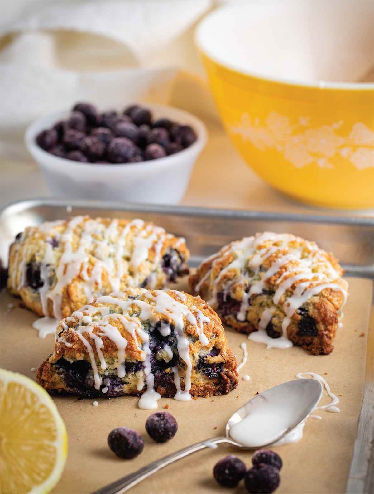 Three glazed blueberry lemon scones on a parchment-lined baking sheet with a lemon half and a spoon on the side.