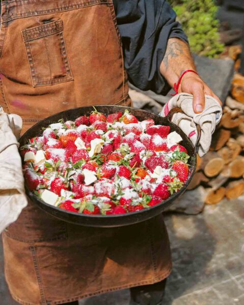A person using a kitchen towel to hold the sides of a large skillet filled with strawberries that are tossed with butter and sugar.