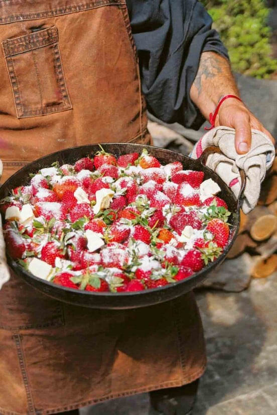 A person using a kitchen towel to hold the sides of a large skillet filled with strawberries that are tossed with butter and sugar.