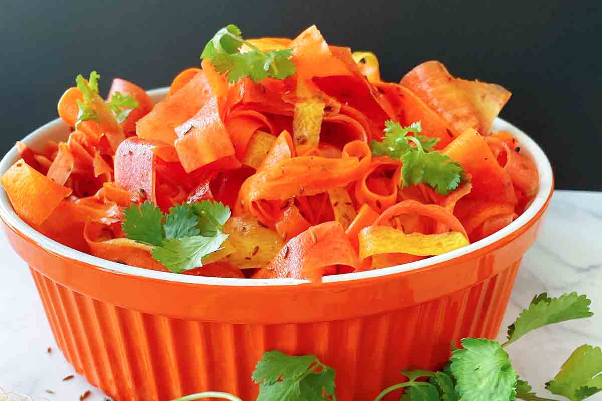 An orange serving bowl filled with carrot ribbon salad, garnished with cilantro leaves and more cilantro leaves in front of the bowl.
