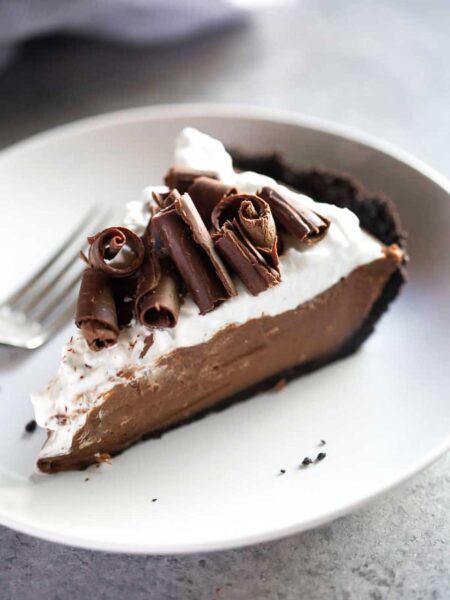 A slice of chocolate cream pie with Oreo crust and chocolate curls on a white plate with a fork resting beside it.