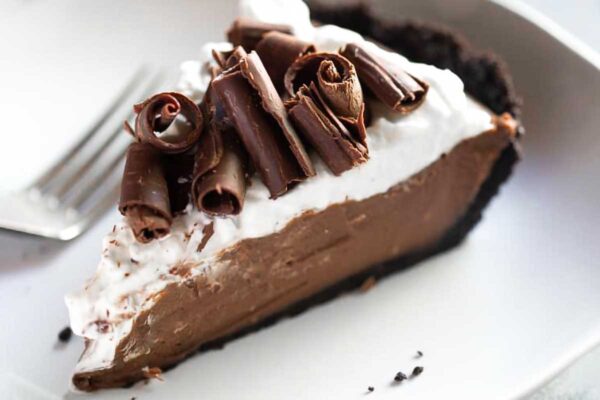 A slice of chocolate cream pie with Oreo crust and chocolate curls on a white plate with a fork resting beside it.