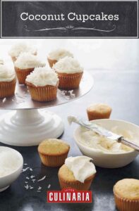 Cake stand with 7 frosted coconut cupcakes, four unfrosted cupcakes on the table, and a bowl of frosting.