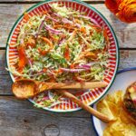 A colorful serving platter filled with coleslaw with cilantro with wooden serving utensils on the side.