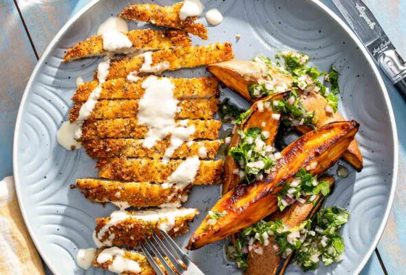 A crispy chicken schnitzel cutlet on a plate with sweet potato wedges and a herb vinaigrette