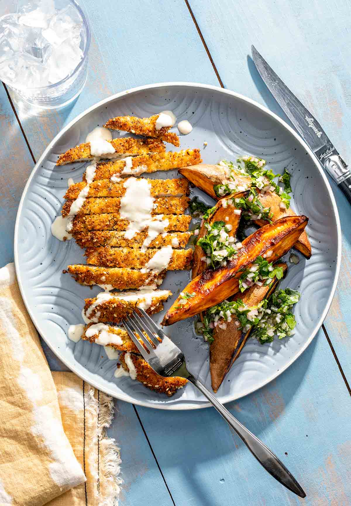 A crispy chicken schnitzel cutlet on a plate with sweet potato wedges and a herb vinaigrette