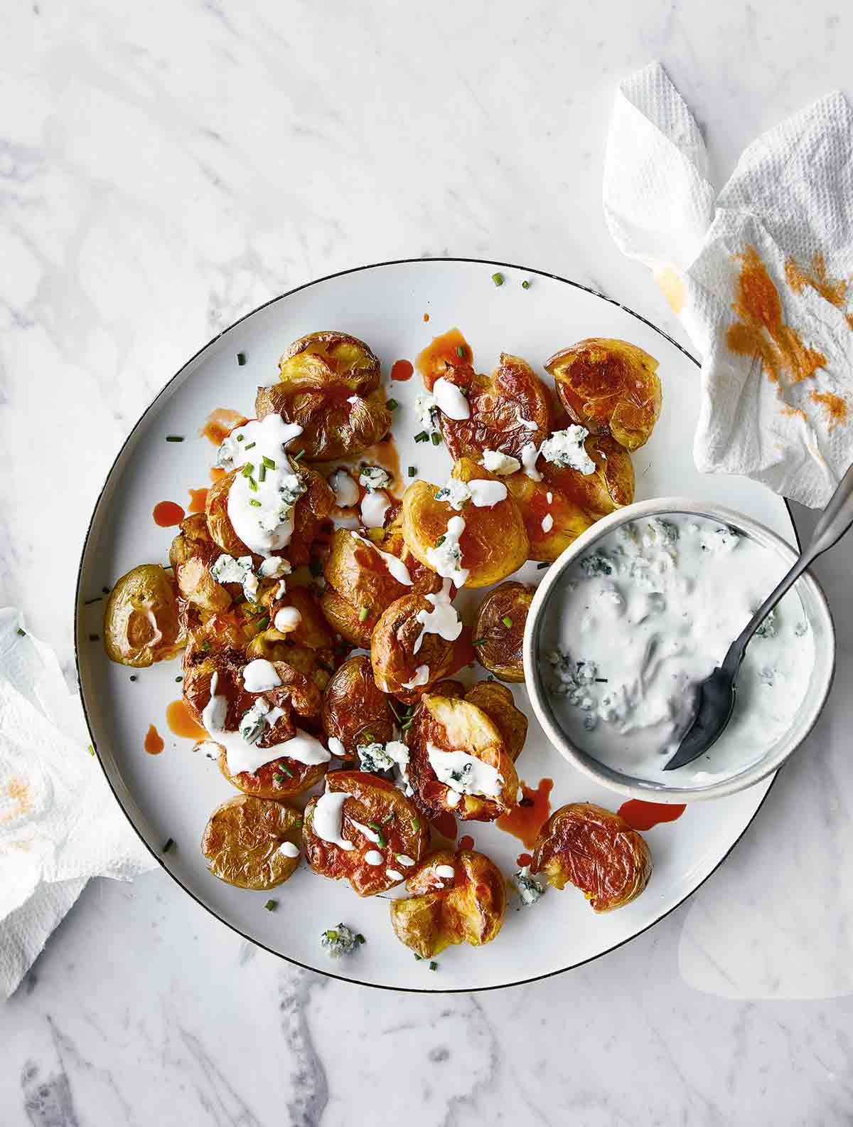 A white plate filled with crispy smashed buffalo potatoes with a bowl of blue cheese sauce on the side and a spoon resting inside.