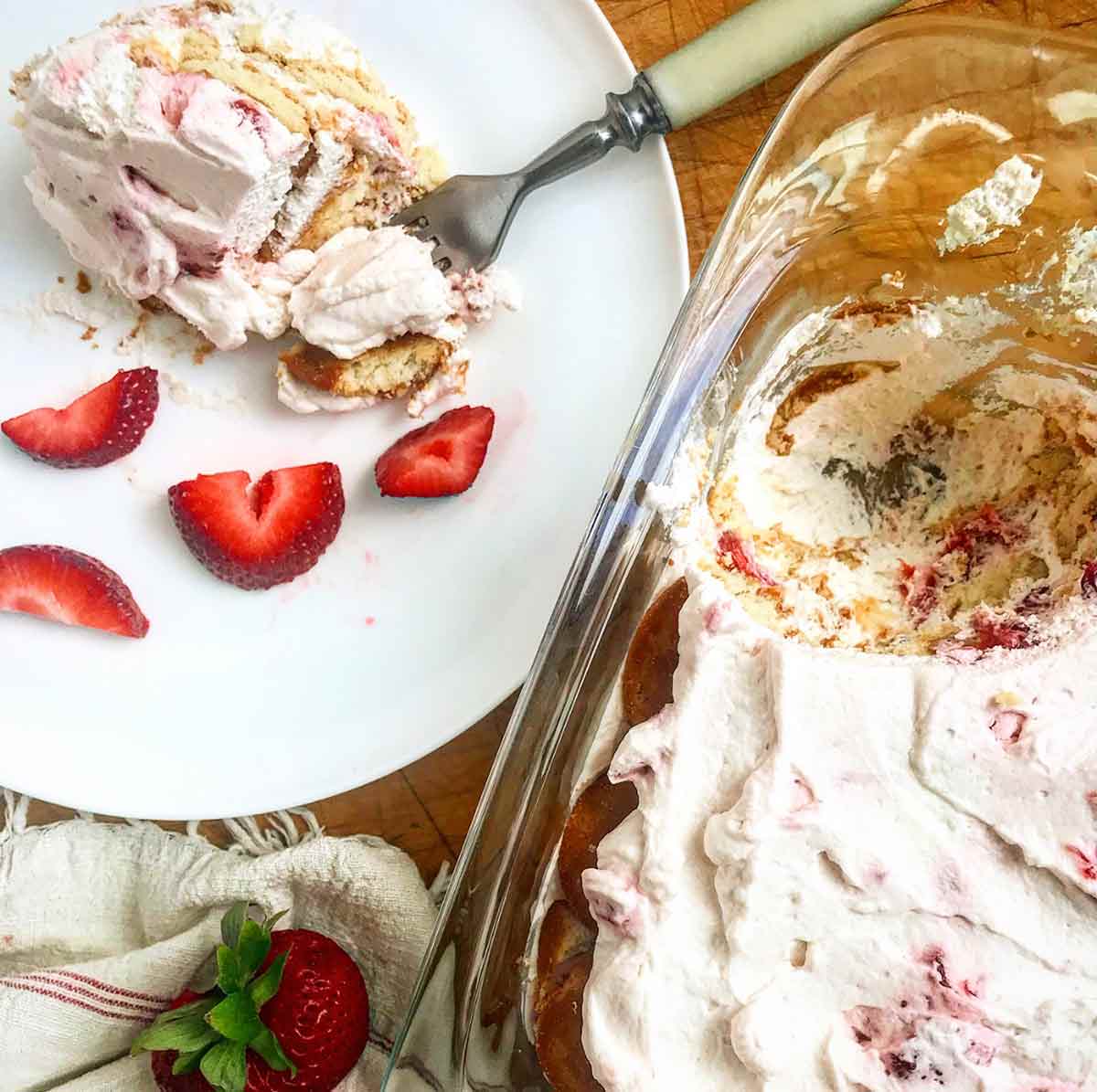 A scoop of easy strawberry icebox cake on a white plate with sliced strawberries and a glass baking dish with the rest of the cake beside it.