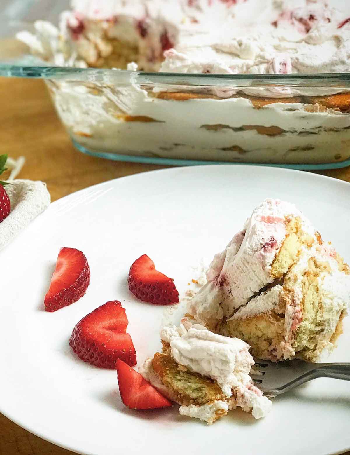 En skopa enkel jordgubbstårta på en vit tallrik med skivade jordgubbar och en ugnsform i glas med resten av kakan bredvid.