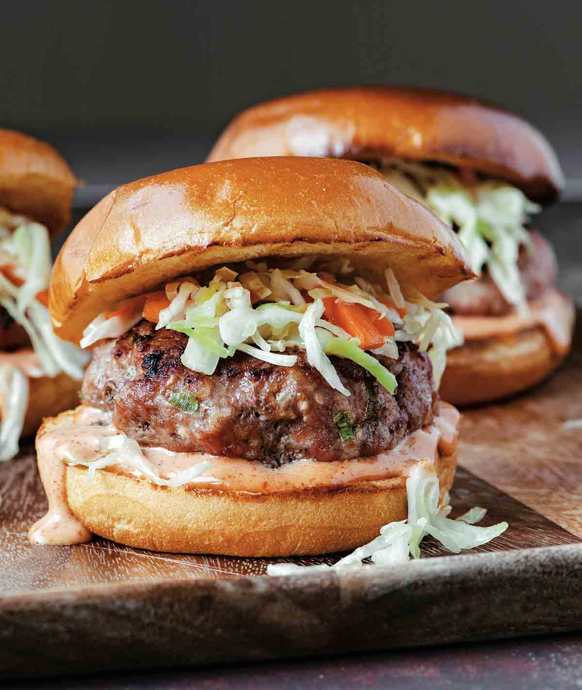 Three grilled pork burgers topped with coleslaw and yum yum sauce on a wooden serving tray.