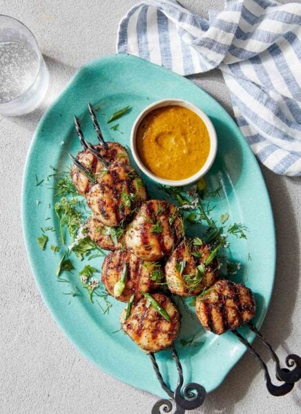 Two grilled scallop kabobs on a blue oval platter with a dish of dipping sauce on the side.