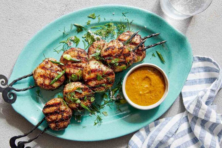 Two grilled scallop kabobs on a blue oval platter with a dish of dipping sauce on the side.